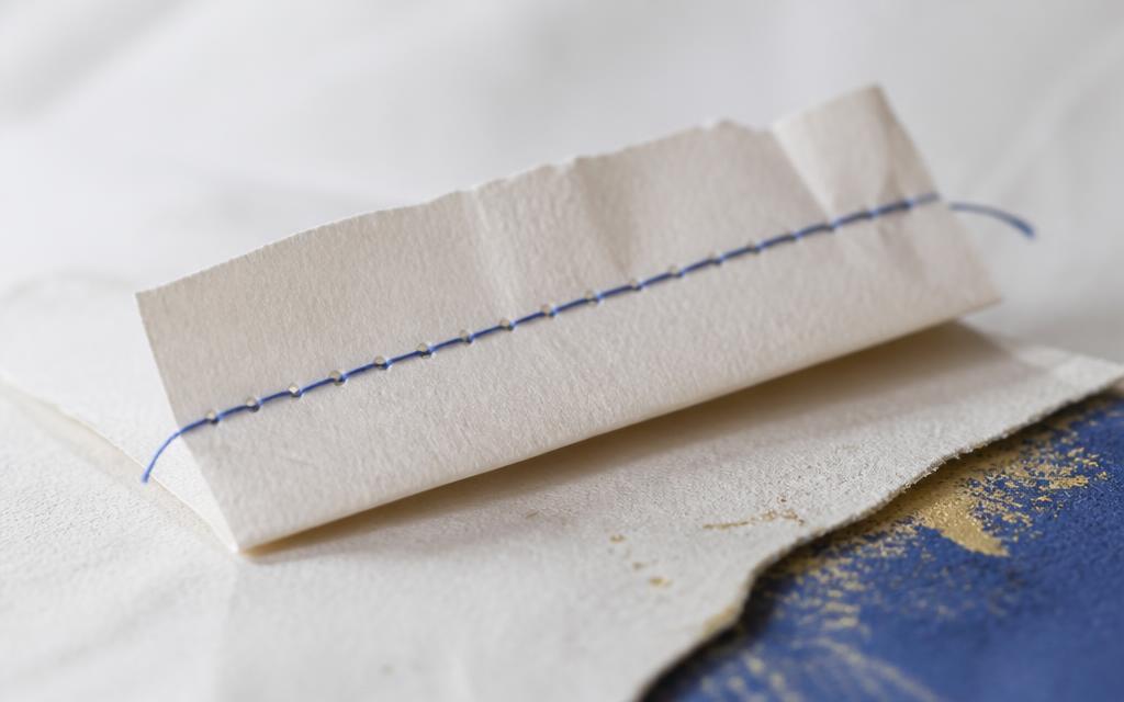 Ein gefalteter Papierstreifen, durch den ein feiner blauer Faden wie eine korrigierte Textzeile gezogen ist.